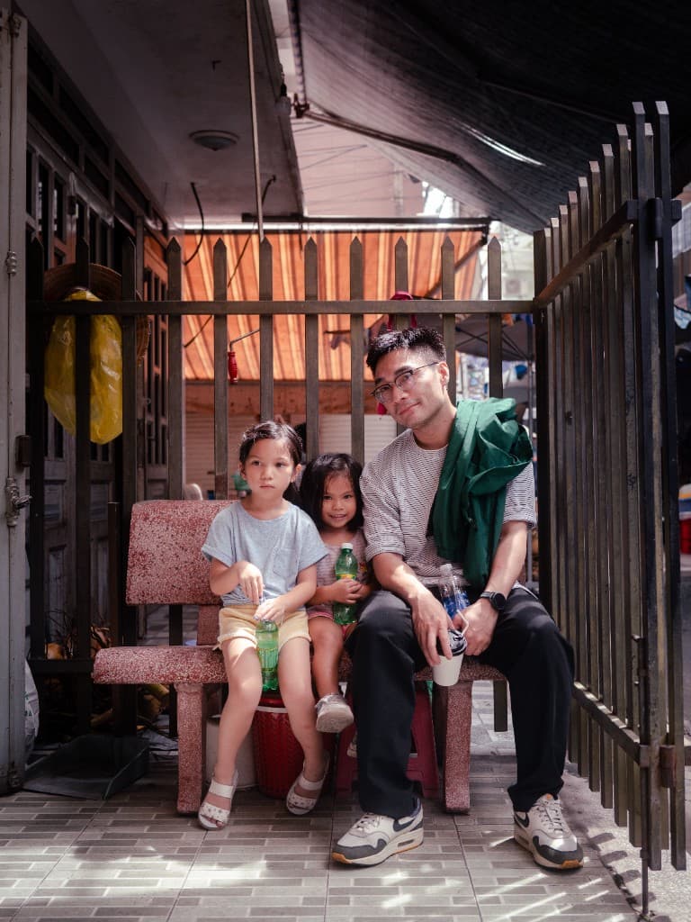 Tan sitting on a stone bench in Biên Hòa with two children, holding a coffee cup.