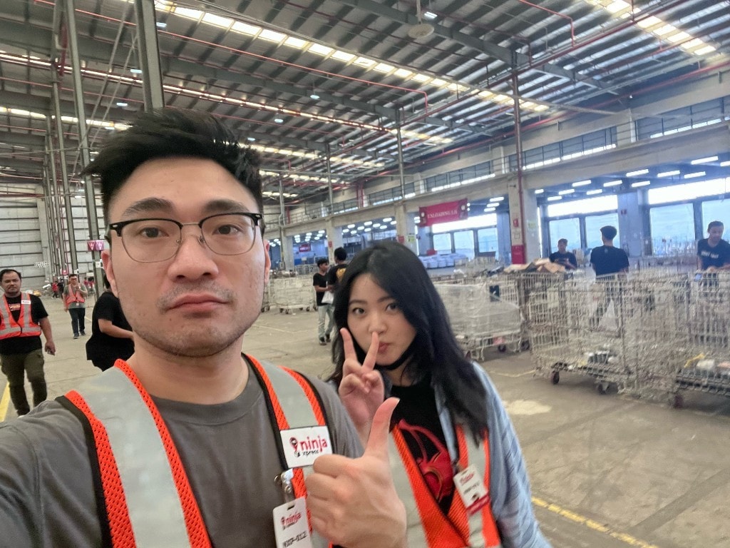 Tan on a Ninja Van warehouse floor in a safety vest, with staff and roll cages in the background.